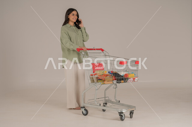 استخدام التقنيات الحديثة, بورتريه امرأة عربية سعودية خليجية ترتدي ثياب كاجوال  تقوم بجر عربة التسوق تحمل بيدها هاتف محمول, قضاء احتياجات المنزل , الإستفادة من العروض والخصومات , الإستمتاع بشراء المستلزمات, صورة الجسم بالطول الكامل, خلفية بيج. 