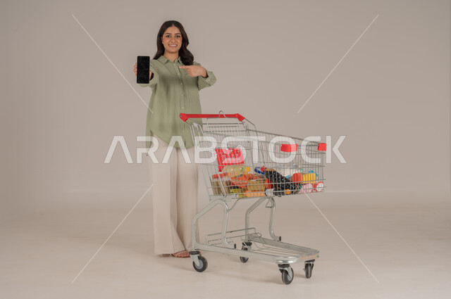 استخدام التقنيات الحديثة, بورتريه امرأة عربية سعودية خليجية ترتدي ثياب كاجوال  تقوم بجر عربة التسوق تشير الى شاشة سوداء في الهاتف محمول, قضاء احتياجات المنزل , الإستفادة من العروض والخصومات , الإستمتاع بشراء المستلزمات, صورة الجسم بالطول الكامل, خلفية بيج. 