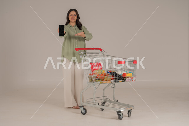استخدام التقنيات الحديثة, بورتريه امرأة عربية سعودية خليجية ترتدي ثياب كاجوال  تقوم بجر عربة التسوق تشير الى شاشة سوداء في الهاتف محمول, قضاء احتياجات المنزل , الإستفادة من العروض والخصومات , الإستمتاع بشراء المستلزمات, صورة الجسم بالطول الكامل, خلفية بيج. 