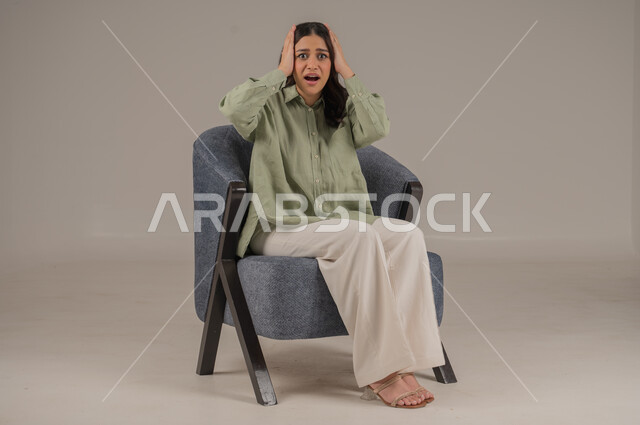 Gestures indicating astonishment at hearing sudden news, shock and wonder at something happening, close-up portrait of a Saudi Arabian Gulf woman in casual wear, hands on her face and looking at the camera with expressions of surprise and fear, beige background