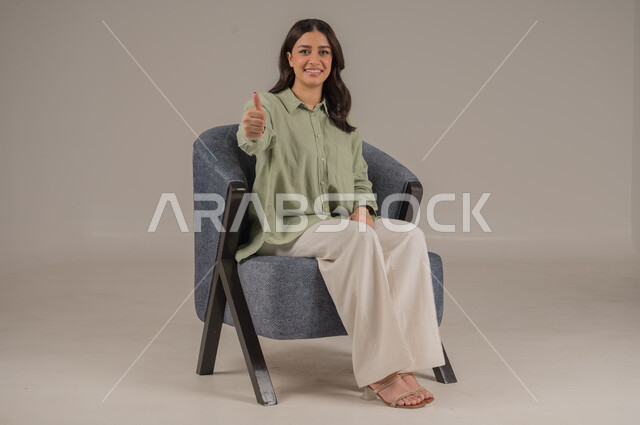 مفهوم القبول والموافقه، تعابير الإعجاب والرضى، بورتريه لامرأه عربيه خليجيه مبتسمة ترتدي الزي الكاجوال ترفع إصبع الابهام دليل على الامتياز والجودة، شابة سعودية انيقة، ايماءات السعادة والسرور، خلفية بيج