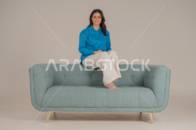 حركات جلوس مختلفة والنظر إلى الكاميرا بإيماءات السعادة والسرور، بورتريه لشابة عربية خليجية سعودية ترتدي الزي الكاجوال تجلس على كنبة مريحة في غرفة المعيشة، الأناقة الاهتمام بالموضة والمظهر الخارجي، الجسم بالطول الكامل، خلفية رمادية