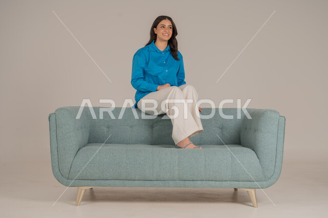 Different sitting movements and looking at the camera with gestures of happiness and pleasure, portrait of a young Saudi Arabian Gulf woman wearing casual clothes sitting on a comfortable sofa in the living room, elegance, interest in fashion and appearance, full body, gray background