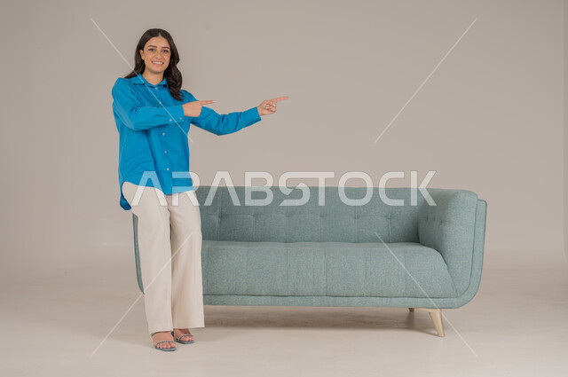 الإشارة بالاصابع نحو اتجاه محدد، النظر إلى الكاميرا بإيماءات السعادة والسرور، بورتريه لشابة عربية خليجية سعودية ترتدي الزي الكاجوال تقف جانب كنبة مريحة تشير باصبيعها السبابة نحو شيء ما، الأناقة والاهتمام بالموضة والمظهر الخارجي، الجسم بالطول الكامل، خلفية رمادية