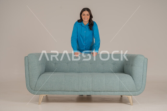 الوقوف برزانة والنظر إلى الكاميرا بإيماءات السعادة والسرور، بورتريه لشابة عربية خليجية سعودية ترتدي الزي الكاجوال تقف خلف كنبة مريحة في غرفة المعيشة، الأناقة الاهتمام بالموضة والمظهر الخارجي، الجسم بالطول الكامل، خلفية رمادية
