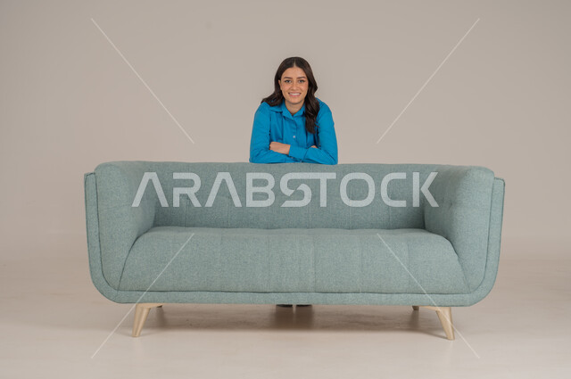 Standing stoically and looking at the camera with gestures of happiness and pleasure, portrait of a young Saudi Arabian Gulf woman in casual wear standing behind a comfortable sofa in the living room, elegance, interest in fashion and appearance, full body, gray background