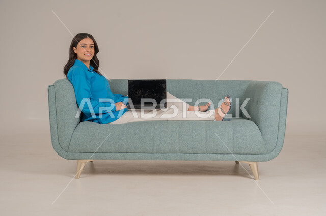 عرض شاشة جوال سوداء فارغة، دمج التقنيات التكنولوجية الحديثة في الحياة اليومية، بورتريه لشابة عربية خليجية سعودية ترتدي الزي الكاجوال تجلس على كنبة مريحة تمسك بيدها جهاز اللابتوب بإيماءات السرور، الاهتمام بالمظهر الخارجي، الجسم بالطول الكامل، خلفية رمادية