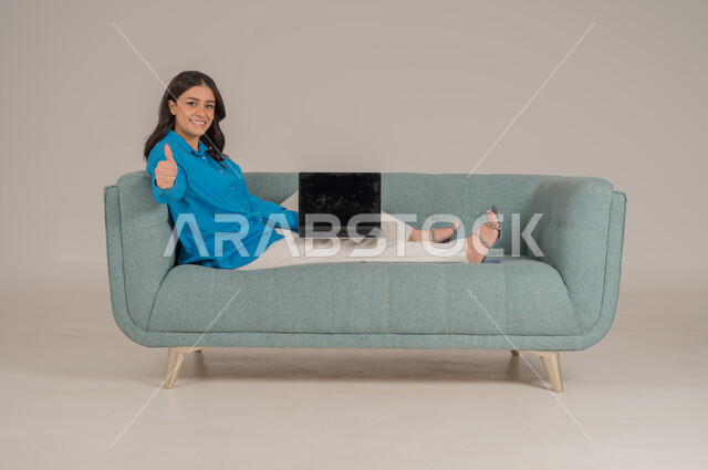 عرض شاشة جوال سوداء فارغة، دمج التقنيات التكنولوجية الحديثة في الحياة اليومية، بورتريه لشابة عربية خليجية سعودية ترتدي الزي الكاجوال تجلس على كنبة مريحة تمسك بيدها جهاز اللابتوب بإيماءات السرور، الاهتمام بالمظهر الخارجي، الجسم بالطول الكامل، خلفية رمادية