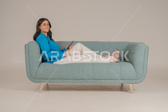 دمج التقنيات التكنولوجية الحديثة في الحياة اليومية، بورتريه لشابة عربية خليجية سعودية ترتدي الزي الكاجوال تجلس على كنبة مريحة تمسك بيدها الهاتف المحمول بإيماءات الاندماج والسعادة، تصفح مواقع الانترنت، الاهتمام بالمظهر الخارجي، الجسم بالطول الكامل، خلفية رمادية