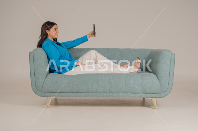 دمج التقنيات التكنولوجية الحديثة في الحياة اليومية، بورتريه لشابة عربية خليجية سعودية ترتدي الزي الكاجوال تجلس على كنبة مريحة تمسك بيدها الهاتف المحمول بإيماءات الاندماج والسعادة، تصفح مواقع الانترنت، الاهتمام بالمظهر الخارجي، الجسم بالطول الكامل، خلفية رمادية
