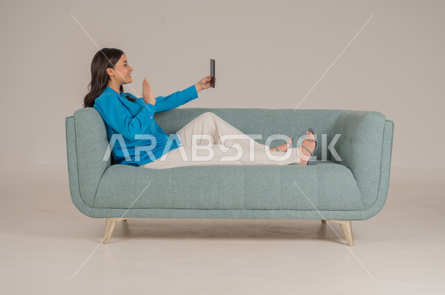 دمج التقنيات التكنولوجية الحديثة في الحياة اليومية، بورتريه لشابة عربية خليجية سعودية ترتدي الزي الكاجوال تجلس على كنبة مريحة تمسك بيدها الهاتف المحمول بإيماءات الاندماج والسعادة، تصفح مواقع الانترنت، الاهتمام بالمظهر الخارجي، الجسم بالطول الكامل، خلفية رمادية