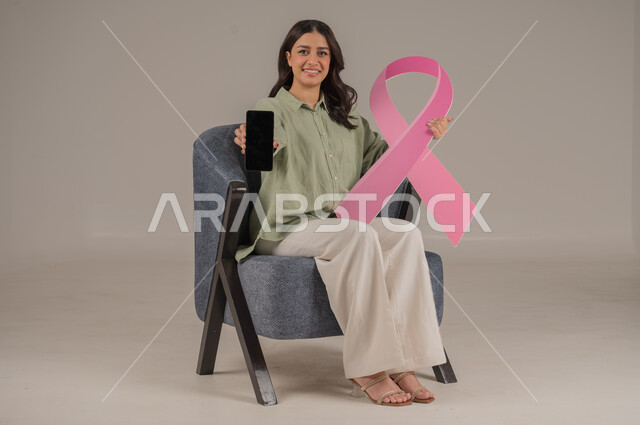الحملة التوعيه بالامراض الخبيثة, استخدام التقنيات الحديثة, بورتريه مقرب لشابة عربية خليجية سعودية مبتسمة ترتدي الزي الكاجوال تمسك بيدها شعار التوعيه للكشف المبكر عن سرطان الثدي تشير الى شاشة سوداء في الهاتف المحمول ، خلفية بيج, محتوى حساس, استخدام حساس