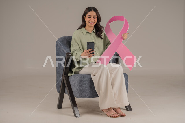 الحملة التوعيه بالامراض الخبيثة, استخدام التقنيات الحديثة, بورتريه مقرب لشابة عربية خليجية سعودية مبتسمة ترتدي الزي الكاجوال تمسك بيدها شعار التوعيه للكشف المبكر عن سرطان الثدي تشير الى شاشة سوداء في الهاتف المحمول ، خلفية بيج, محتوى حساس, استخدام حساس
