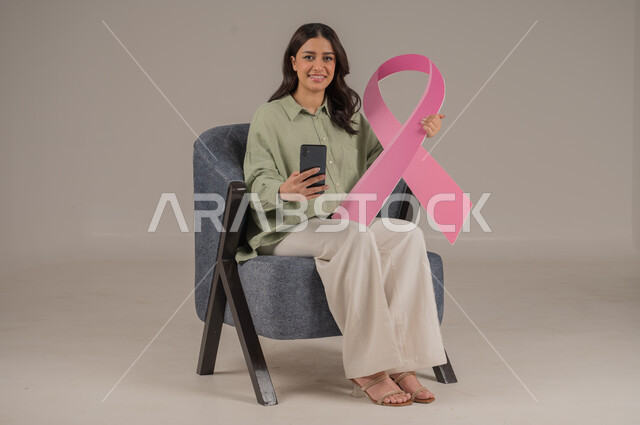 الحملة التوعيه بالامراض الخبيثة, استخدام التقنيات الحديثة, بورتريه مقرب لشابة عربية خليجية سعودية مبتسمة ترتدي الزي الكاجوال تمسك بيدها شعار التوعيه للكشف المبكر عن سرطان الثدي تشير الى شاشة سوداء في الهاتف المحمول ، خلفية بيج, محتوى حساس, استخدام حساس