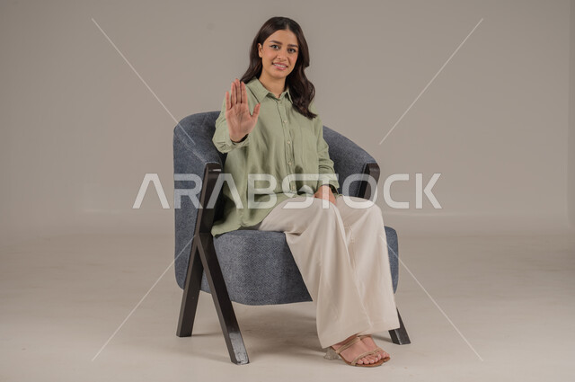 الصمت والتوقف عن الكلام، إيماءات يدين تدل على الأمر بالتوقف عن فعل شيء ما و النهي عنه, بورتريه لشابة عربية خليجية سعودية مبتسمة ترتدي الزي الكاجوال تجلس على الكرسي ترفع كفها للأعلى, خلفية بيج