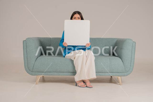 لوحات توضيحية ووسائل تعليمية، العروض والإعلانات التسويقية, بورتريه لإمرأة عربية خليجية سعودية تجلس على كنبة تمسك بيديها لوحة بيضاء مستطيلة فارغة، شابة سعودية تنظر إلى الكاميرا بتعابير السعادة والسرور، موك اب مستطيل أبيض, خلفية رمادية