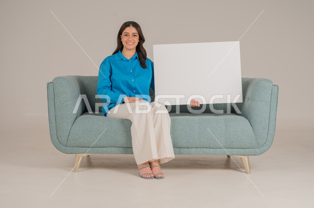 لوحات توضيحية ووسائل تعليمية، العروض والإعلانات التسويقية, بورتريه لإمرأة عربية خليجية سعودية تجلس على كنبة تمسك بيديها لوحة بيضاء مستطيلة فارغة، شابة سعودية تنظر إلى الكاميرا بتعابير السعادة والسرور، موك اب مستطيل أبيض, خلفية رمادية