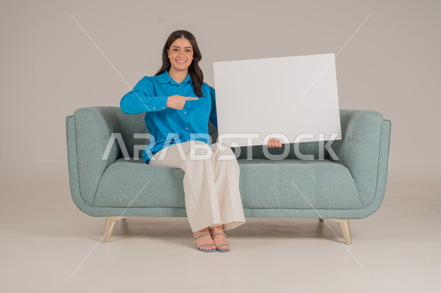 لوحات توضيحية ووسائل تعليمية، العروض والإعلانات التسويقية, بورتريه لإمرأة عربية خليجية سعودية تجلس على كنبة تمسك بيديها لوحة بيضاء مستطيلة فارغة، شابة سعودية تنظر إلى الكاميرا بتعابير السعادة والسرور، موك اب مستطيل أبيض, خلفية رمادية