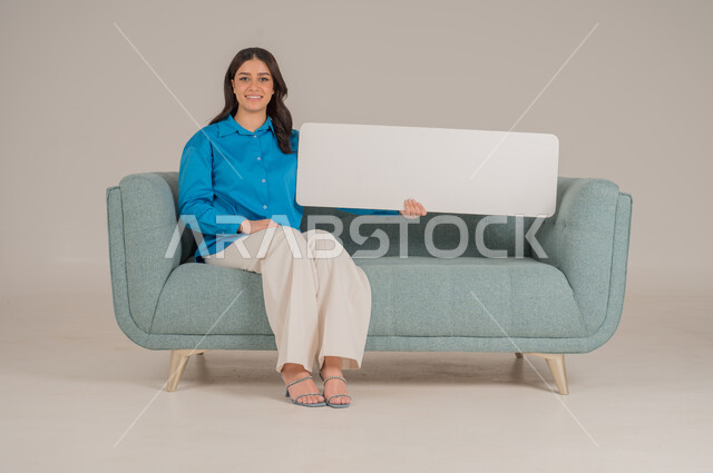 لوحات توضيحية ووسائل تعليمية، العروض والإعلانات التسويقية, بورتريه لإمرأة عربية خليجية سعودية تجلس على كنبة تمسك بيديها لوحة بيضاء مستطيلة فارغة، شابة سعودية تنظر إلى الكاميرا بتعابير السعادة والسرور، موك اب مستطيل أبيض, خلفية رمادية