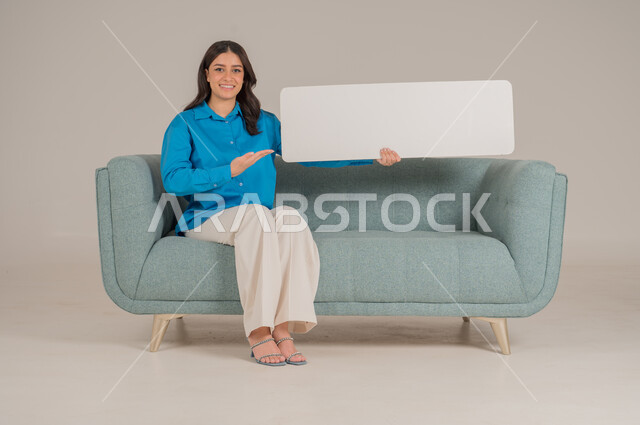 لوحات توضيحية ووسائل تعليمية، العروض والإعلانات التسويقية, بورتريه لإمرأة عربية خليجية سعودية تجلس على كنبة تمسك بيديها لوحة بيضاء مستطيلة فارغة، شابة سعودية تنظر إلى الكاميرا بتعابير السعادة والسرور، موك اب مستطيل أبيض, خلفية رمادية