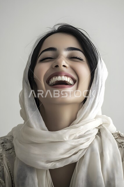 تعابير الأنوثة الناعمة, الاهتمام بالمظهر الخارجي, الوقوف بإيماءات السرور والسعادة, بورتريه لإمرأة عربية خليجية تضحك ترتدي الحجاب, الاستمتاع بقضاء وقت ممتع, خلفية رمادية