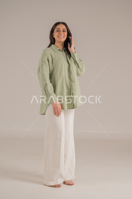 قضاء اوقات ممتعة بتبادل الاخبار، تعابير الفرح عند سماع الاخبار السارة، استخدام الأجهزة التقنية الحديثة، بورتريه لإمرأة عربية سعودية مبتسمة ترتدي الزي الكاجوال تقوم بإجراء مكالمة هاتفية، الاهتمام بالأناقة والمظهر الخارجي، الجسم بالطول الكامل، خلفية بيج