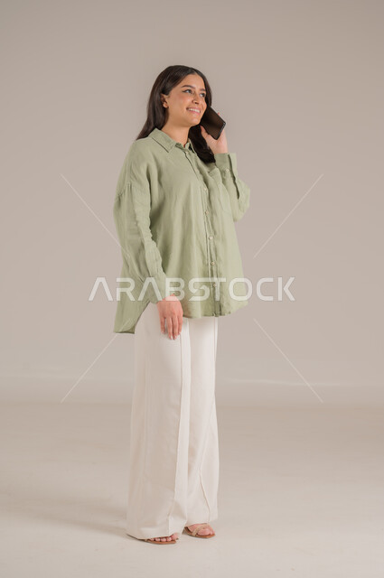 قضاء اوقات ممتعة بتبادل الاخبار، تعابير الفرح عند سماع الاخبار السارة، استخدام الأجهزة التقنية الحديثة، بورتريه لإمرأة عربية سعودية مبتسمة ترتدي الزي الكاجوال تقوم بإجراء مكالمة هاتفية، الاهتمام بالأناقة والمظهر الخارجي، الجسم بالطول الكامل، خلفية بيج