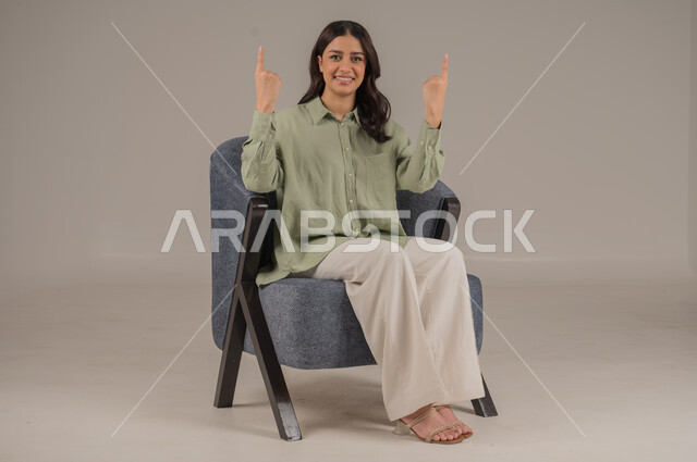 الإشارة بالاصابع نحو اتجاه محدد، النظر إلى الكاميرا بإيماءات السعادة والسرور، بورتريه لشابة عربية خليجية سعودية ترتدي الزي الكاجوال تجلس على كنبة مريحة تشير باصبيعها السبابة نحو الأعلى، الأناقة والاهتمام بالموضة والمظهر الخارجي، خلفية بيج