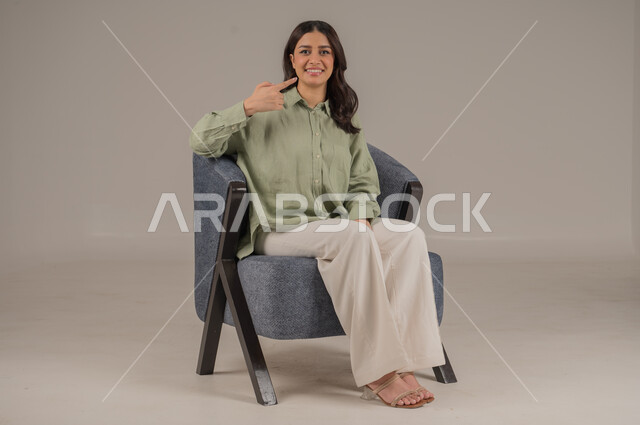 النظر إلى الكاميرا بإيماءات السرور والفرح، أهمية المحافظة على صحة الفم والأسنان من التسوس، بورتريه لشابة عربية خليجية سعودية مبتسمة ترتدي الزي الكاجوال تجلس على كنبة مريحة تشير باصبع السبابة إلى أسنانها، خلفية بيج