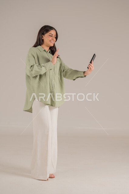 قضاء اوقات ممتعة بتبادل الاخبار، تعابير الفرح عند سماع الاخبار السارة، استخدام الأجهزة التقنية الحديثة، بورتريه لإمرأة عربية سعودية مبتسمة ترتدي الزي الكاجوال تقوم بإجراء مكالمة فيديو، الاهتمام بالأناقة والمظهر الخارجي، الجسم بالطول الكامل، خلفية بيج