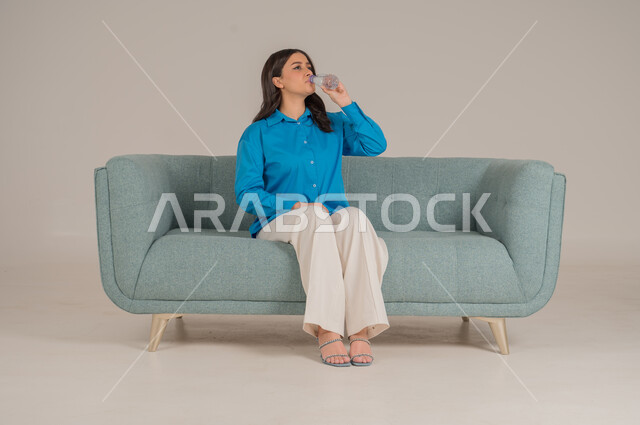 الجلوس والنظر إلى الكاميرا بإيماءات السعادة والسرور، بورتريه لشابة عربية خليجية سعودية ترتدي الزي الكاجوال تجلس على كنبة مريحة في غرفة المعيشة، الأناقة الاهتمام بالموضة والمظهر الخارجي، الجسم بالطول الكامل، خلفية رمادية