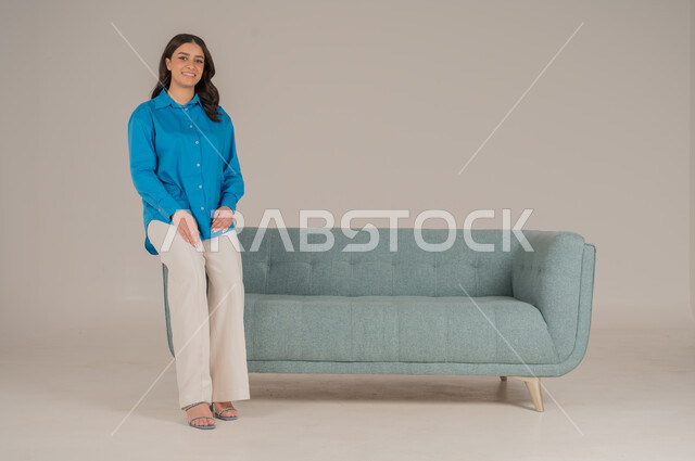 الجلوس والنظر إلى الكاميرا بإيماءات السعادة والسرور، بورتريه لشابة عربية خليجية سعودية ترتدي الزي الكاجوال تجلس على كنبة مريحة في غرفة المعيشة، الأناقة الاهتمام بالموضة والمظهر الخارجي، الجسم بالطول الكامل، خلفية رمادية