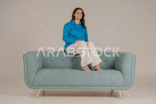الجلوس والنظر إلى الكاميرا بإيماءات السعادة والسرور، بورتريه لشابة عربية خليجية سعودية ترتدي الزي الكاجوال تجلس على كنبة مريحة في غرفة المعيشة، الأناقة الاهتمام بالموضة والمظهر الخارجي، الجسم بالطول الكامل، خلفية رمادية