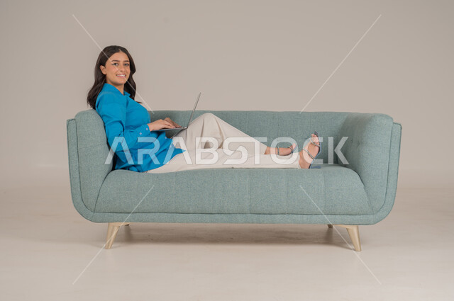 دمج التقنيات التكنولوجية الحديثة في الحياة اليومية، بورتريه لشابة عربية خليجية سعودية ترتدي الزي الكاجوال تجلس على كنبة مريحة تمسك بيدها جهاز اللابتوب بإيماءات السرور، الاهتمام بالمظهر الخارجي، الجسم بالطول الكامل، خلفية رمادية