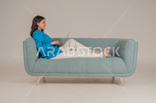 دمج التقنيات التكنولوجية الحديثة في الحياة اليومية، بورتريه لشابة عربية خليجية سعودية ترتدي الزي الكاجوال تجلس على كنبة مريحة تمسك بيدها جهاز اللابتوب بإيماءات السرور، الاهتمام بالمظهر الخارجي، الجسم بالطول الكامل، خلفية رمادية