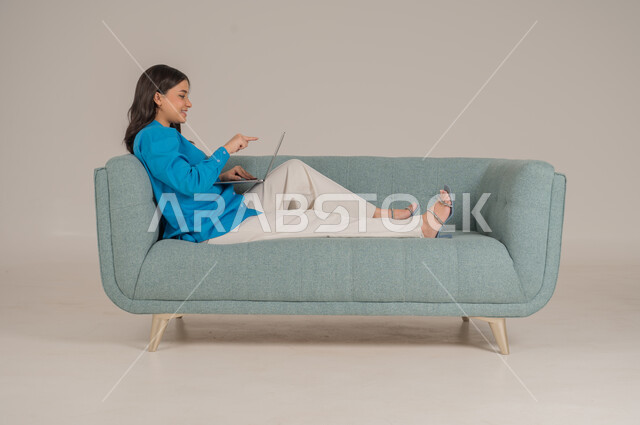 دمج التقنيات التكنولوجية الحديثة في الحياة اليومية، بورتريه لشابة عربية خليجية سعودية ترتدي الزي الكاجوال تجلس على كنبة مريحة تمسك بيدها جهاز اللابتوب بإيماءات السرور، الاهتمام بالمظهر الخارجي، الجسم بالطول الكامل، خلفية رمادية