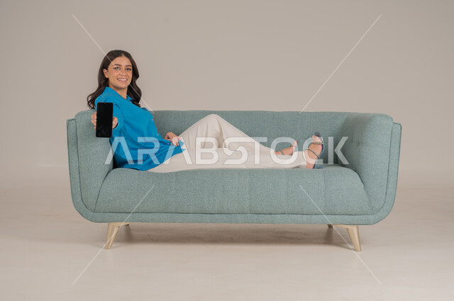 Empty black mobile screen, integration of modern technology into daily life, portrait of a young Saudi Arabian Gulf woman in casual wear sitting on a comfortable sofa holding a mobile phone with gestures of pleasure, concern for appearance, full body, gray background