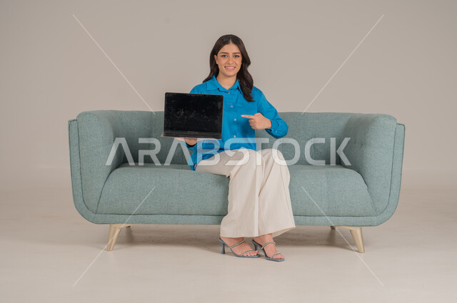 Empty black mobile screen, integration of modern technology into daily life, portrait of a young Saudi Arabian Gulf woman in casual wear sitting on a comfortable sofa holding a laptop with gestures of pleasure, concern for appearance, full body, gray background