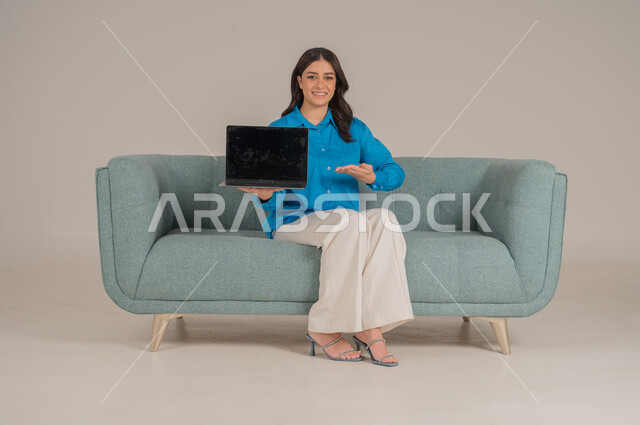 عرض شاشة جوال سوداء فارغة، دمج التقنيات التكنولوجية الحديثة في الحياة اليومية، بورتريه لشابة عربية خليجية سعودية ترتدي الزي الكاجوال تجلس على كنبة مريحة تمسك بيدها اللابتوب بإيماءات السرور، الاهتمام بالمظهر الخارجي، الجسم بالطول الكامل، خلفية رمادية