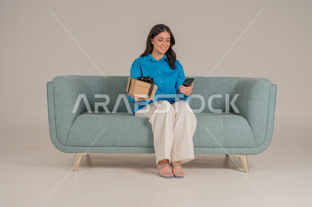 The concept of gifting and appreciation, giving gifts on holidays and occasions to express love, portrait of a young Saudi Arabian Gulf woman wearing casual clothes sitting on a sofa holding an elegantly wrapped gift box and a mobile phone and looking at the camera with gestures of pleasure and happiness, full body, gray background