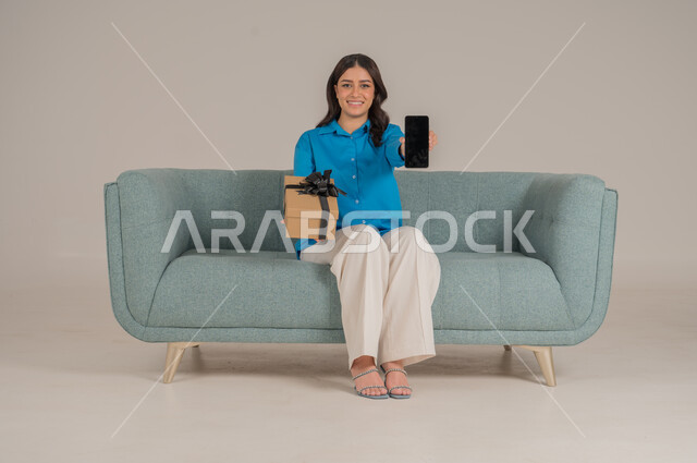 The concept of gifting and appreciation, giving gifts on holidays and occasions to express love, portrait of a young Saudi Arabian Gulf woman wearing casual clothes sitting on a sofa holding an elegantly wrapped gift box and a mobile phone and looking at the camera with gestures of pleasure and happiness, full body, gray background