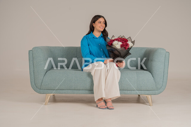 The concept of gifting and appreciation, giving gifts on holidays and occasions to express love, portrait of a young Saudi Arabian Gulf woman wearing casual clothes sitting on a sofa holding a bouquet of roses wrapped elegantly and looking at the camera with gestures of pleasure and happiness, full body, gray background