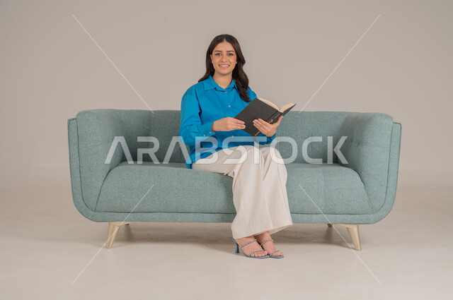 Spending time reading, learning and educating yourself, portrait of a Saudi Arabian Gulf woman wearing casual clothes holding a book in her hands, enjoying reading books, stories and novels, completing daily duties, self-learning culture, gray background