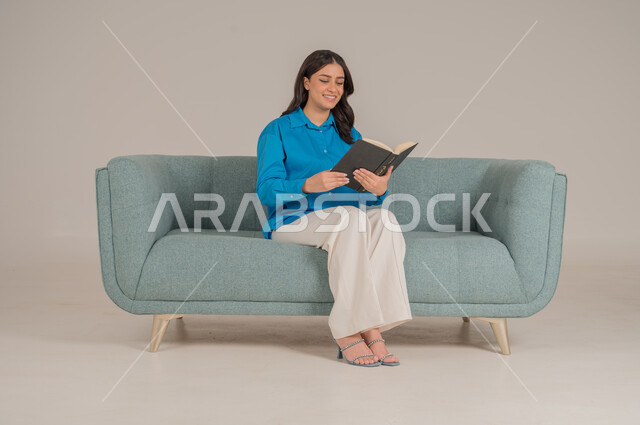 Spending time reading, learning and educating yourself, portrait of a Saudi Arabian Gulf woman wearing casual clothes holding a book in her hands, enjoying reading books, stories and novels, completing daily duties, self-learning culture, gray background