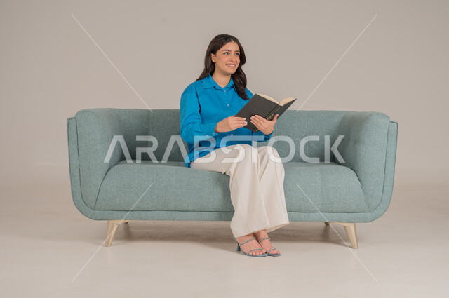 Spending time reading, learning and educating yourself, portrait of a Saudi Arabian Gulf woman wearing casual clothes holding a book in her hands, enjoying reading books, stories and novels, completing daily duties, self-learning culture, gray background