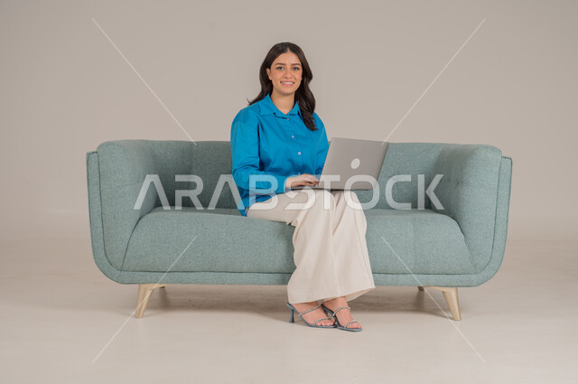 دمج التقنيات التكنولوجية الحديثة في الحياة اليومية، بورتريه لشابة عربية خليجية سعودية ترتدي الزي الكاجوال تجلس على كنبة مريحة تمسك بيدها جهاز اللابتوب بإيماءات السرور، الاهتمام بالمظهر الخارجي، الجسم بالطول الكامل، خلفية رمادية