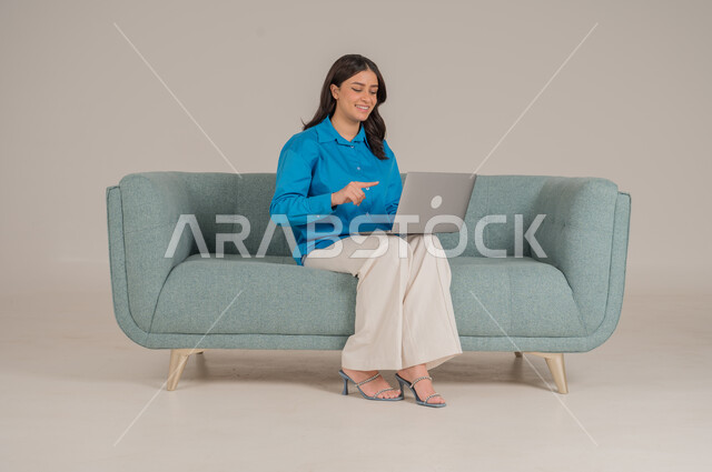 دمج التقنيات التكنولوجية الحديثة في الحياة اليومية، بورتريه لشابة عربية خليجية سعودية ترتدي الزي الكاجوال تجلس على كنبة مريحة تمسك بيدها جهاز اللابتوب بإيماءات السرور، الاهتمام بالمظهر الخارجي، الجسم بالطول الكامل، خلفية رمادية