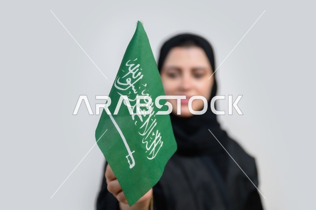 بورتريه لسيدة عربية خليجية سعودية تحمل بيدها علم المملكة العربية السعودية بإيماءات تدل على السعادة ، اليوم الوطني السعودي ، الإحتفال بالعيد الوطني ، خلفية بيضاء