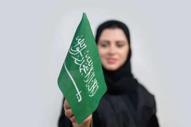 بورتريه لسيدة عربية خليجية سعودية تحمل بيدها علم المملكة العربية السعودية بإيماءات تدل على السعادة ، اليوم الوطني السعودي ، الإحتفال بالعيد الوطني ، خلفية بيضاء