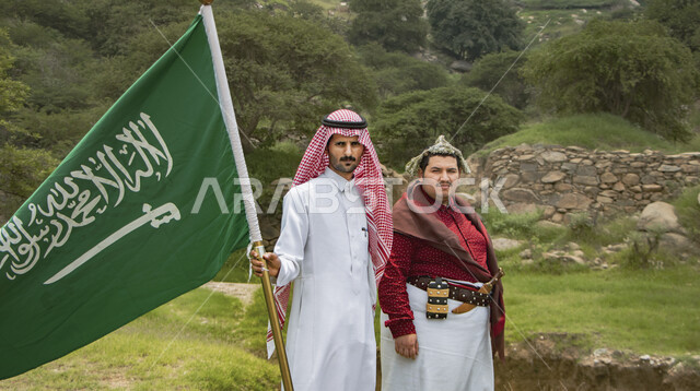 صورة لرجل عربي خليجي سعودي  يرتدي الزي السعودي الخاص بمنطقة جازان بجانبه رجل خليجي سعودي يرتدي الزي السعودي التقليدي ، يحمل بيده علم المملكة العربية السعودية ،ذكرى اليوم الوطني السعودي ، الاحتفال بالعيد الوطني السعودي